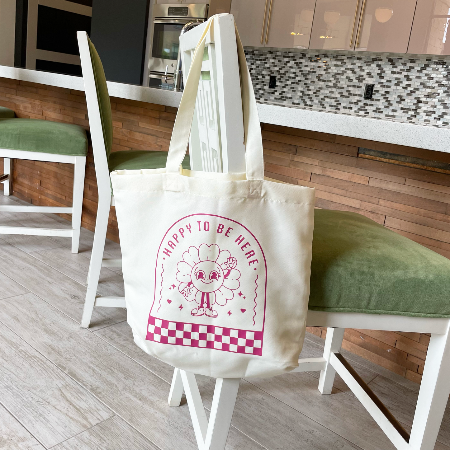 tote bag with design saying happy to be here with a happy flower waving hanging from a chair