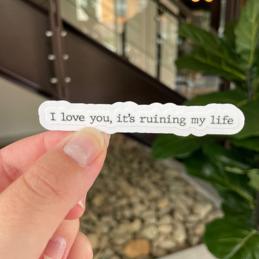 hand holding a sticker in typewriter font saying "I love you, it's ruining my life"