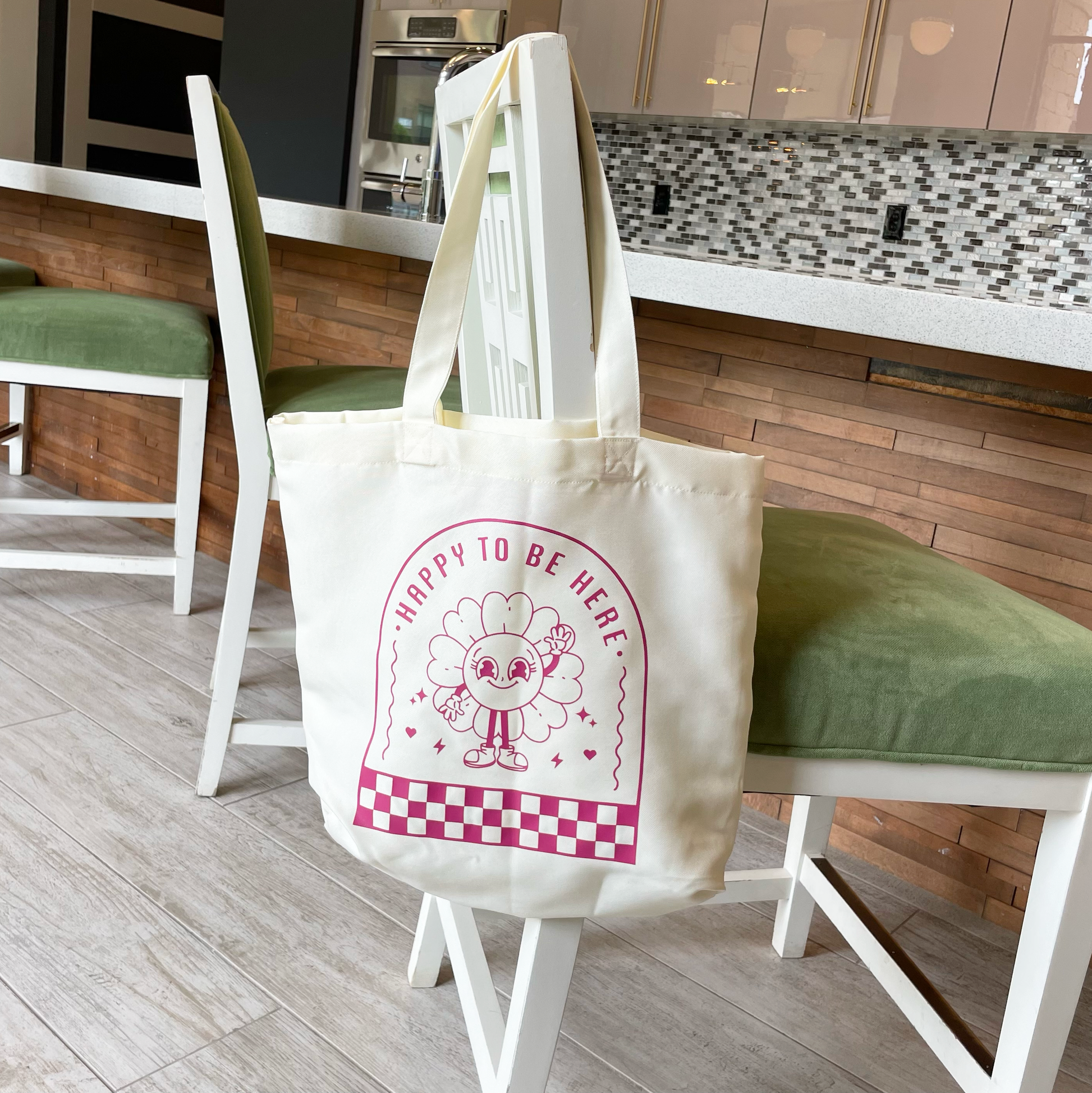 tote bag with design saying happy to be here with a happy flower waving hanging from a chair