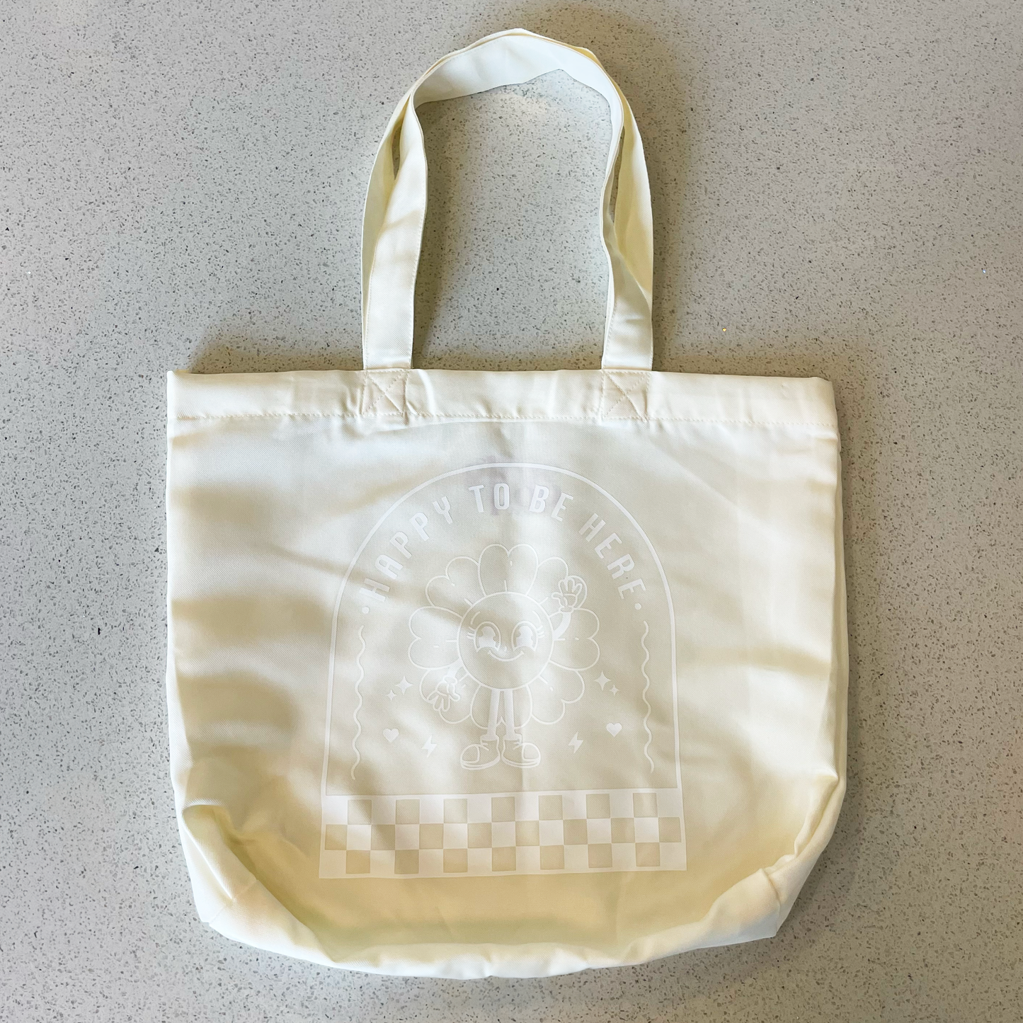 tote bag with design saying happy to be here with a happy flower waving in white