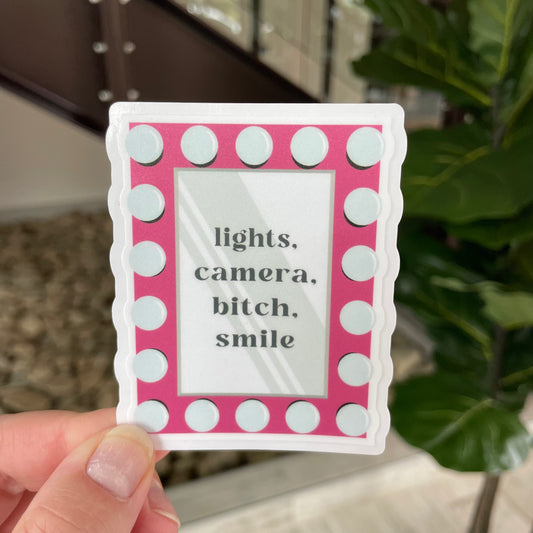 hand holding sticker of a vanity mirror that says lights camera bitch smile