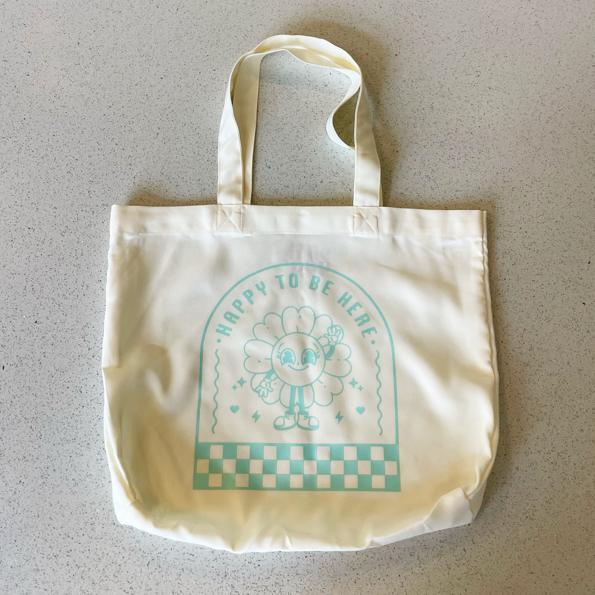 tote bag with design saying happy to be here with a happy flower waving in mint green